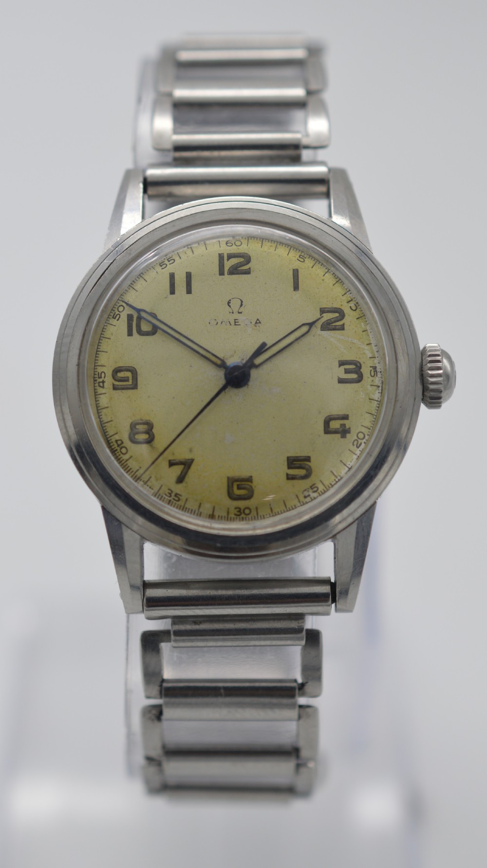 A 1944 Omega military-style wristwatch featuring a patinated cream dial, black Arabic numerals, dark hands, a stainless steel case, and a flexible metal bracelet.