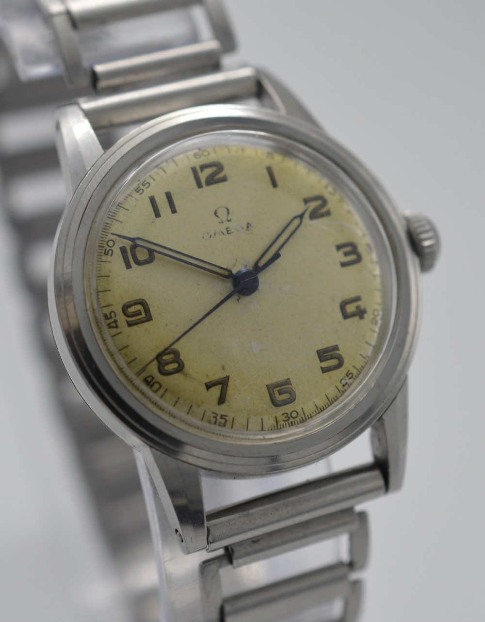 A 1944 Omega military-style wristwatch featuring a patinated cream dial, black Arabic numerals, dark hands, a stainless steel case, and a flexible metal bracelet. View IV