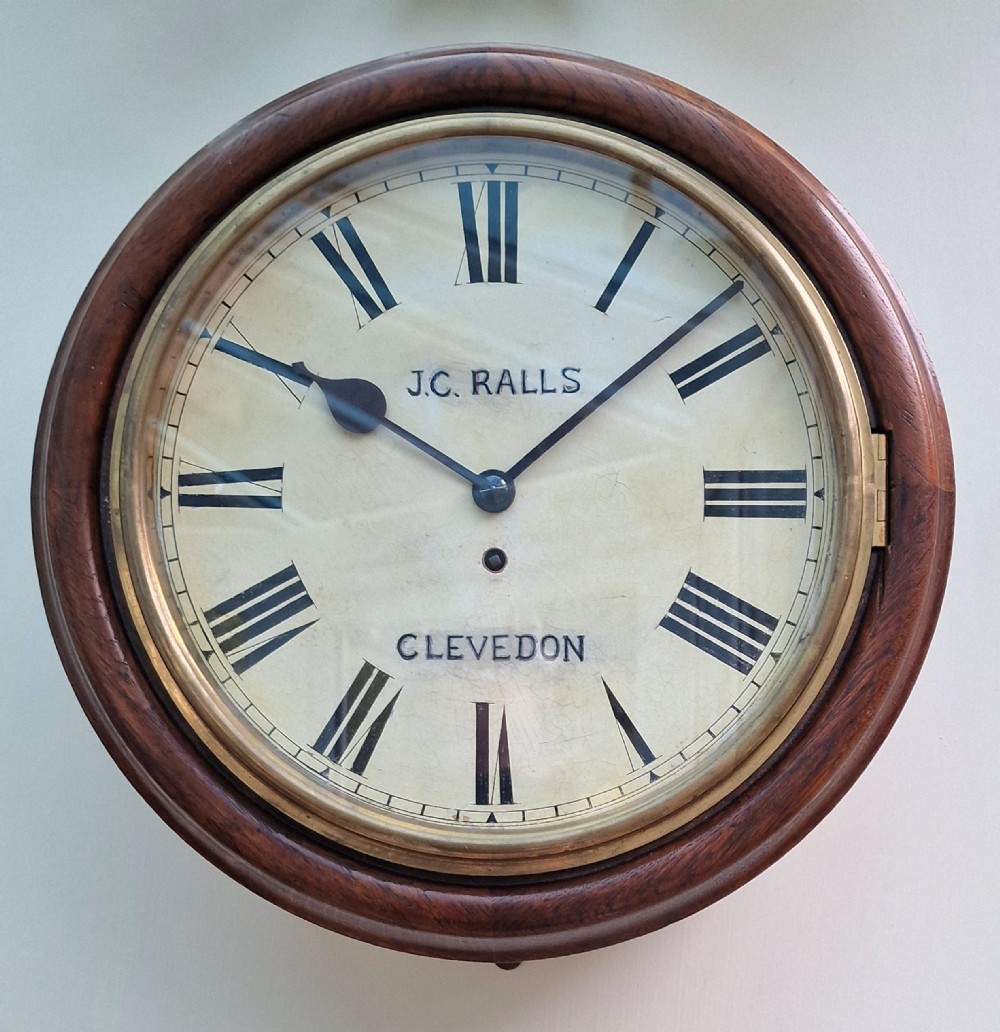 A circular antique wall clock featuring a mahogany wooden surround, hinged brass bezel, and off-white painted dial inscribed with J.C. Ralls Clevedon.