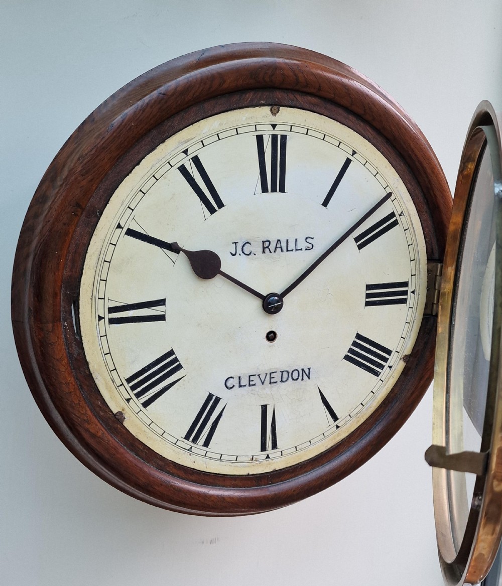 A circular antique wall clock featuring a mahogany wooden surround, hinged brass bezel, and off-white painted dial inscribed with J.C. Ralls Clevedon. View V