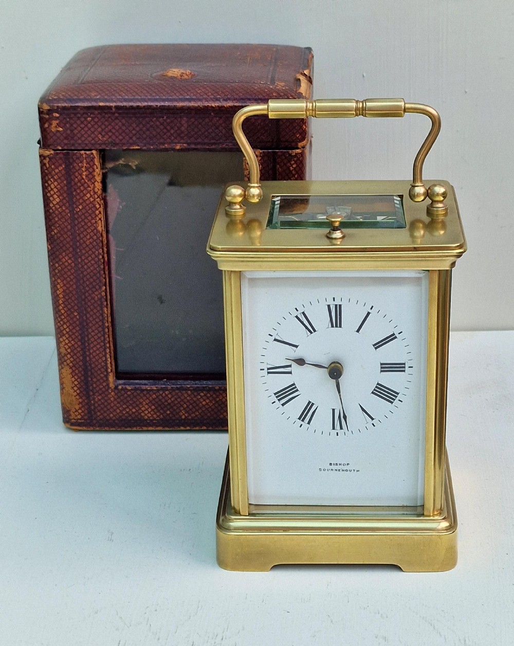 An antique brass repeating carriage clock featuring a white enamel dial with Roman numerals, bevelled glass panels, and a hinged top handle.