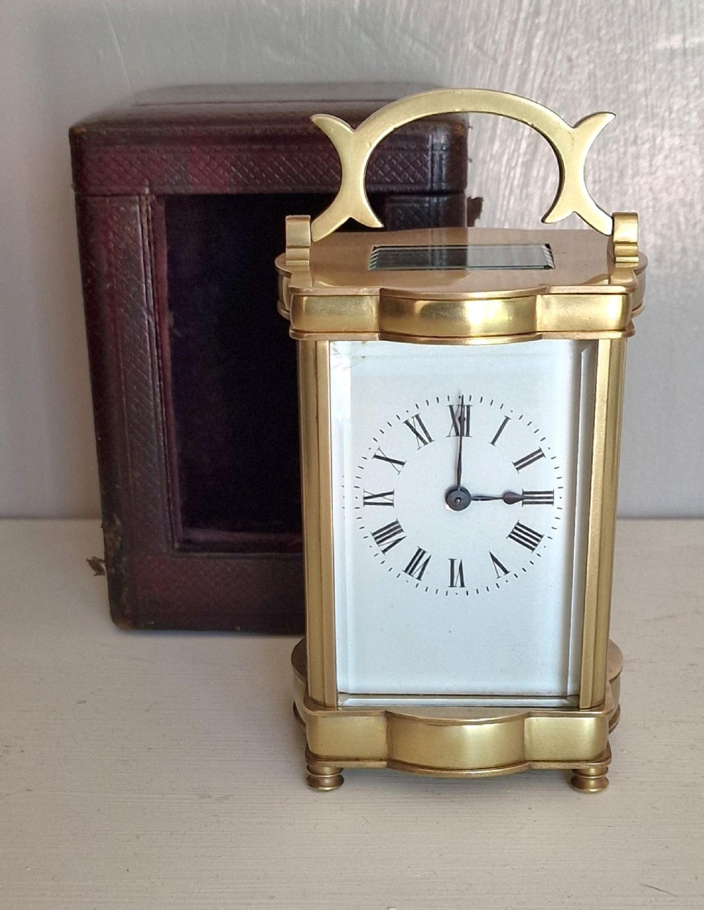 A brass serpentine carriage clock featuring a white enamel dial with Roman numerals, bevelled glass panels, and an arched carrying handle.