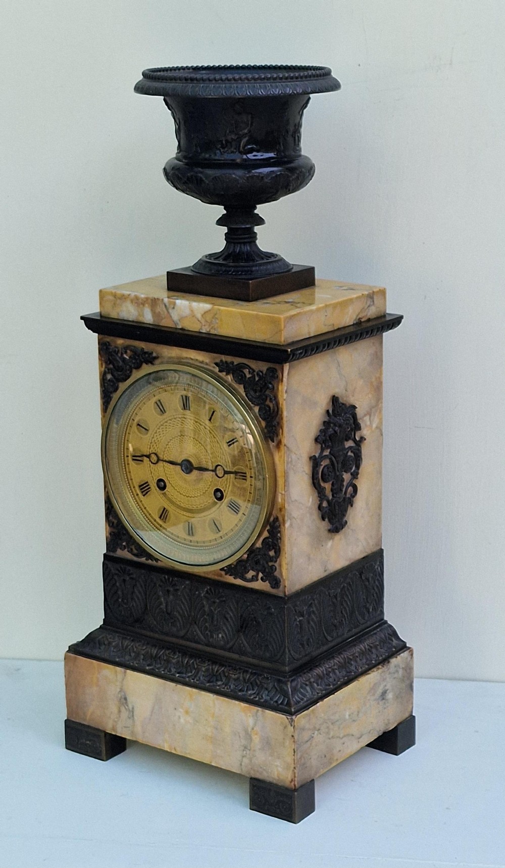 A rectangular Sienna marble mantel clock featuring a dark patinated bronze classical urn finial, a gilt dial with Roman numerals, and detailed bronze corner mounts.