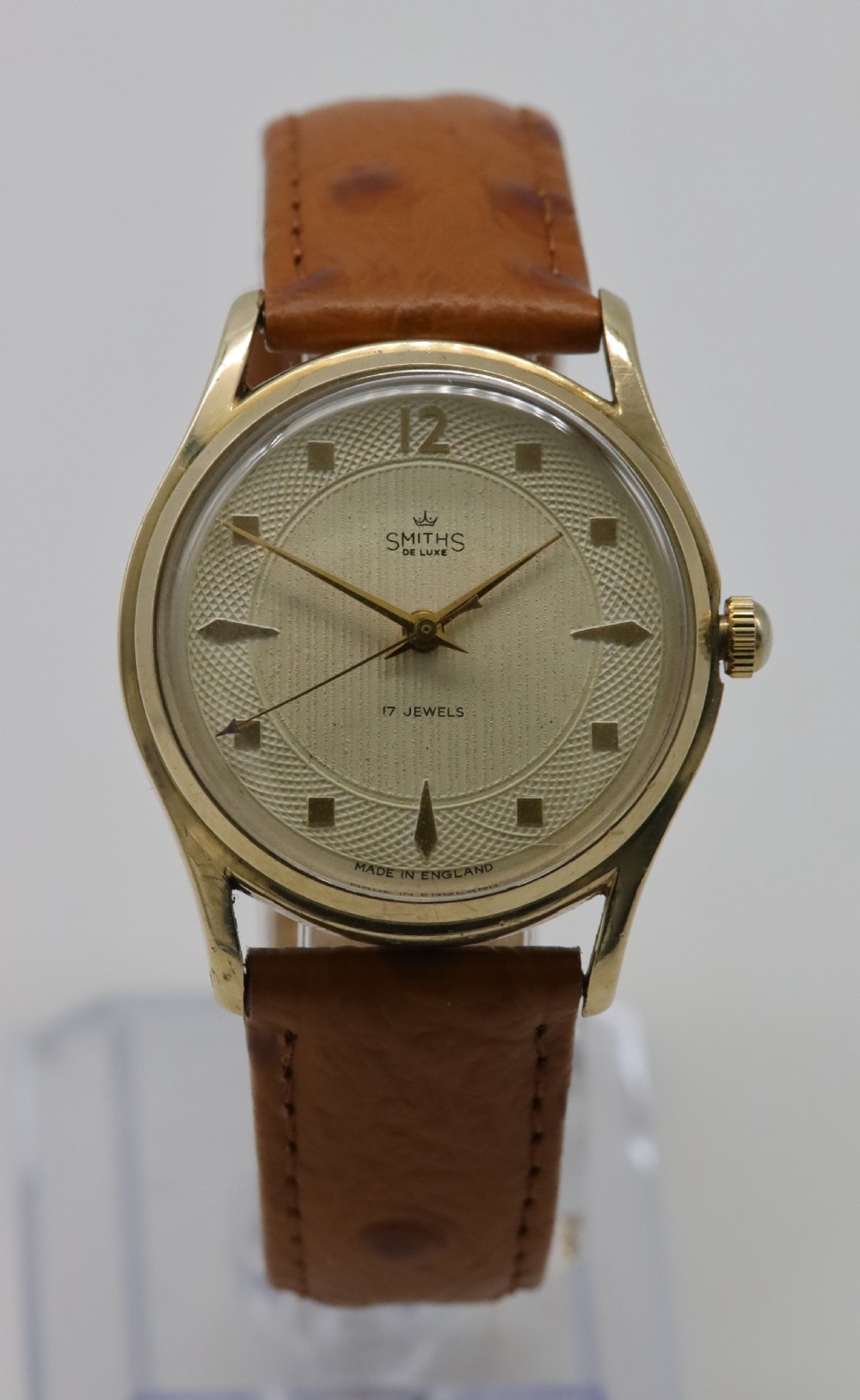 A vintage mid-century Smiths De Luxe wristwatch featuring a 9ct gold round case, textured cream dial with gold hour markers, and a tan leather strap.