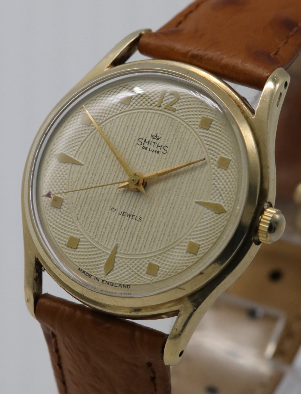 A vintage mid-century Smiths De Luxe wristwatch featuring a 9ct gold round case, textured cream dial with gold hour markers, and a tan leather strap. View II