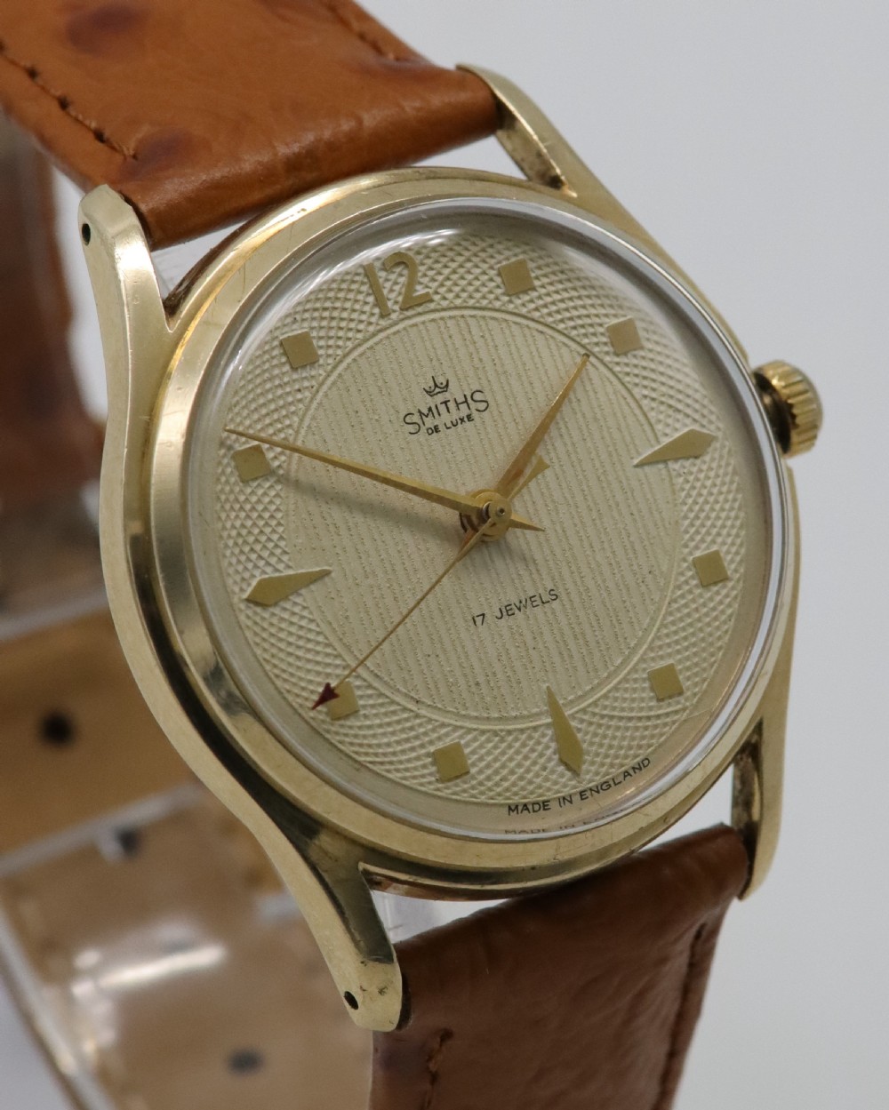 A vintage mid-century Smiths De Luxe wristwatch featuring a 9ct gold round case, textured cream dial with gold hour markers, and a tan leather strap. View IV