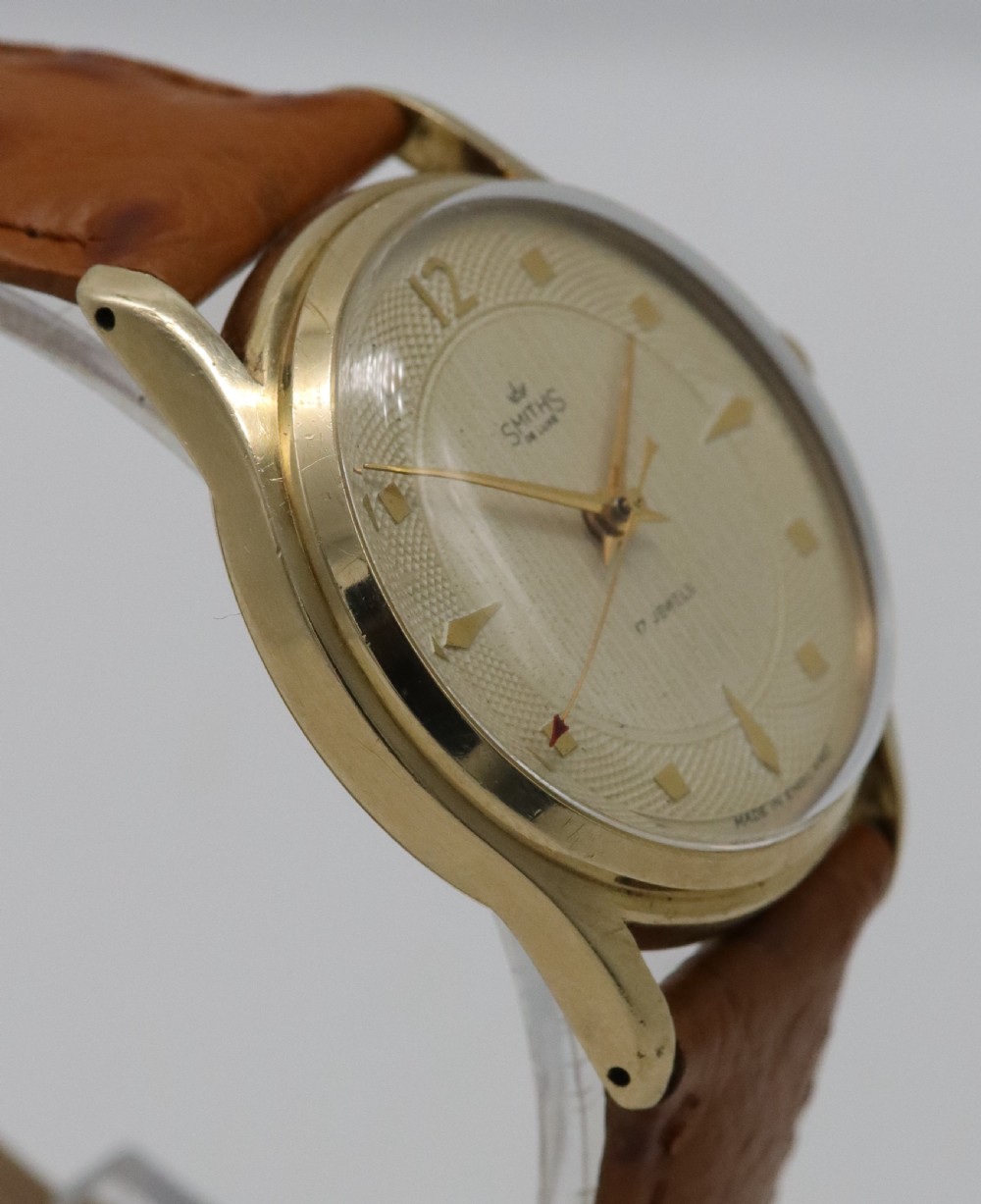A vintage mid-century Smiths De Luxe wristwatch featuring a 9ct gold round case, textured cream dial with gold hour markers, and a tan leather strap. View V