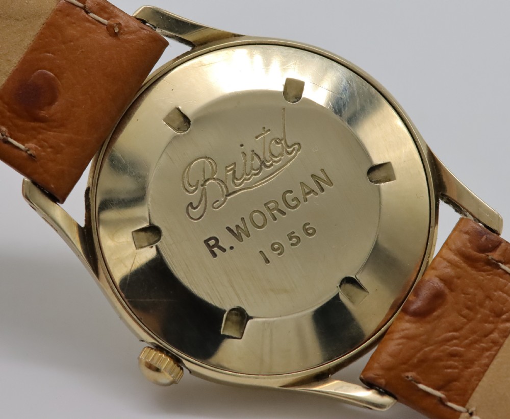 A vintage mid-century Smiths De Luxe wristwatch featuring a 9ct gold round case, textured cream dial with gold hour markers, and a tan leather strap. View VI