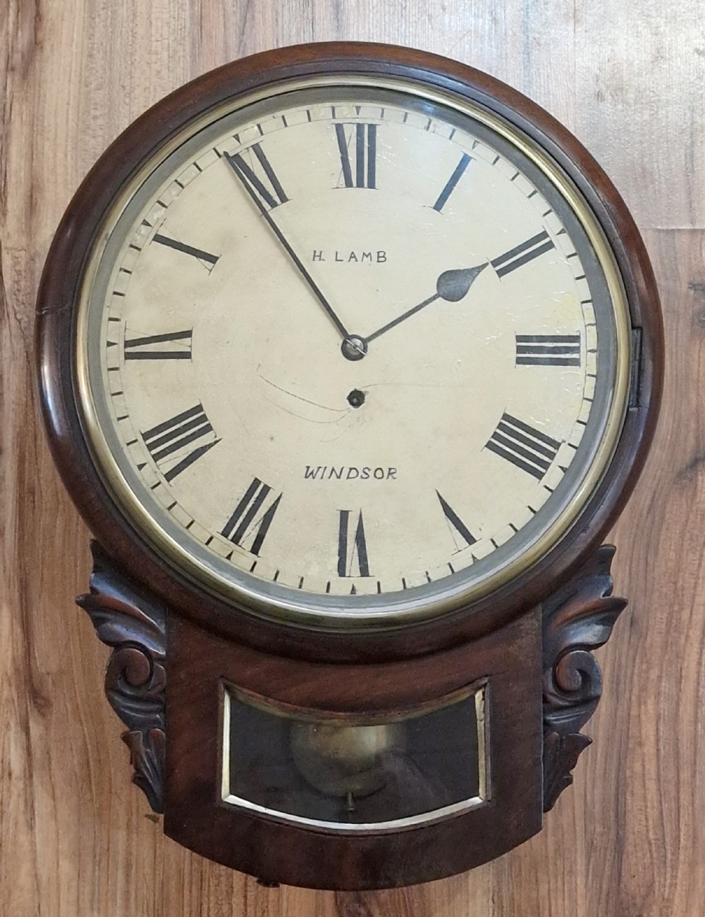 An antique mahogany drop-dial wall clock featuring a painted dial with Roman numerals signed H. Lamb Windsor, a circular brass bezel, carved scroll ears, and a lower pendulum aperture.