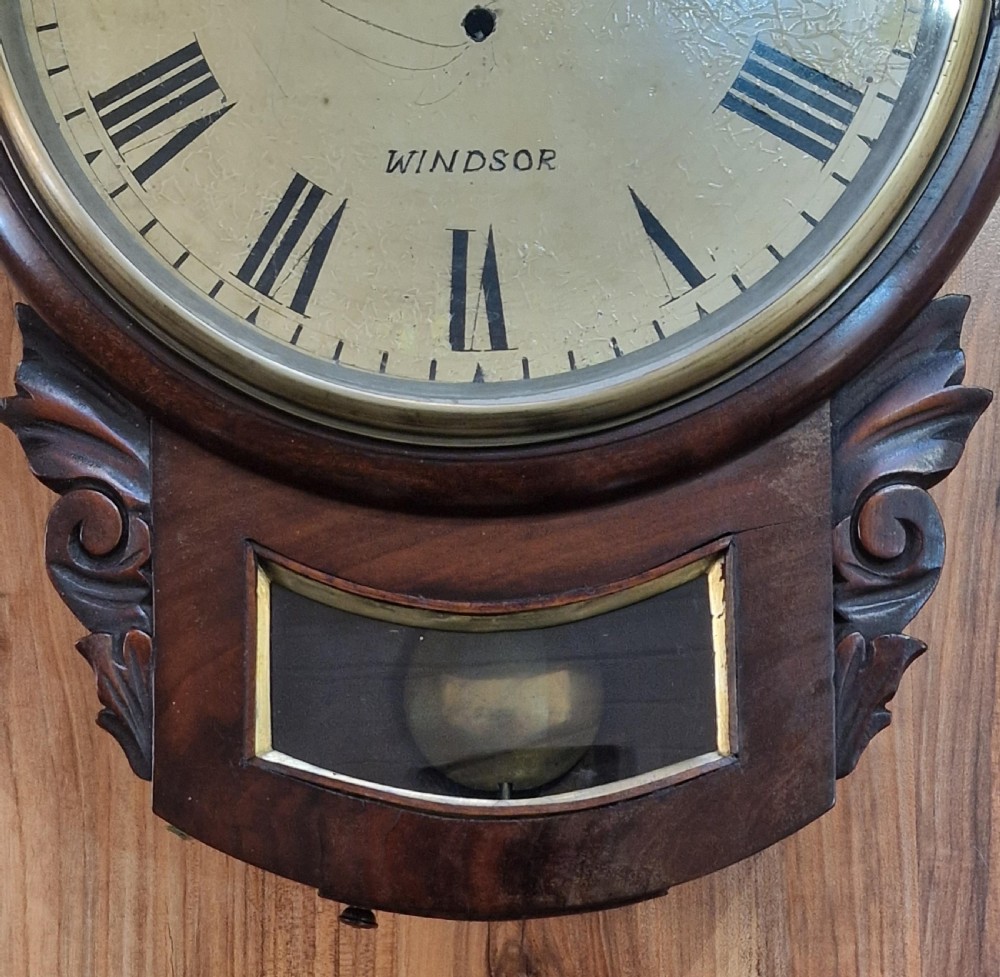 An antique mahogany drop-dial wall clock featuring a painted dial with Roman numerals signed H. Lamb Windsor, a circular brass bezel, carved scroll ears, and a lower pendulum aperture. View II