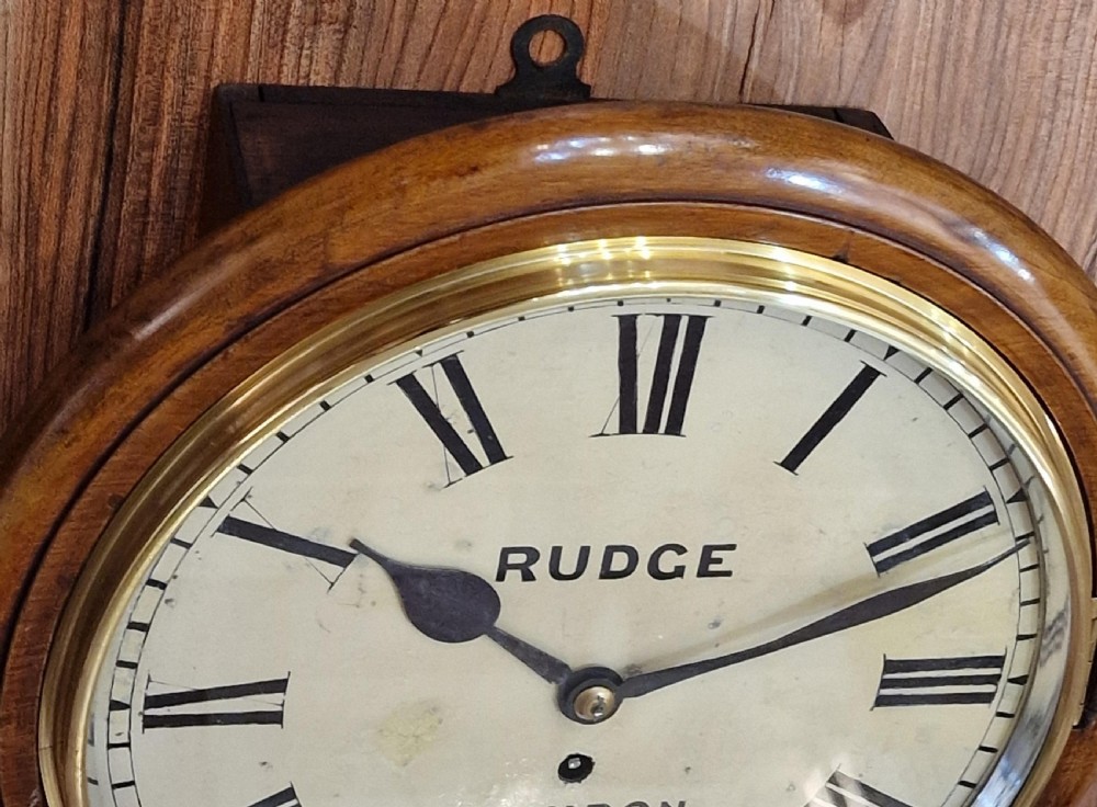 A circular Victorian mahogany fusee dial wall clock featuring a white face with Roman numerals, a brass bezel, and black spade hands, marked Rudge Swindon. View V
