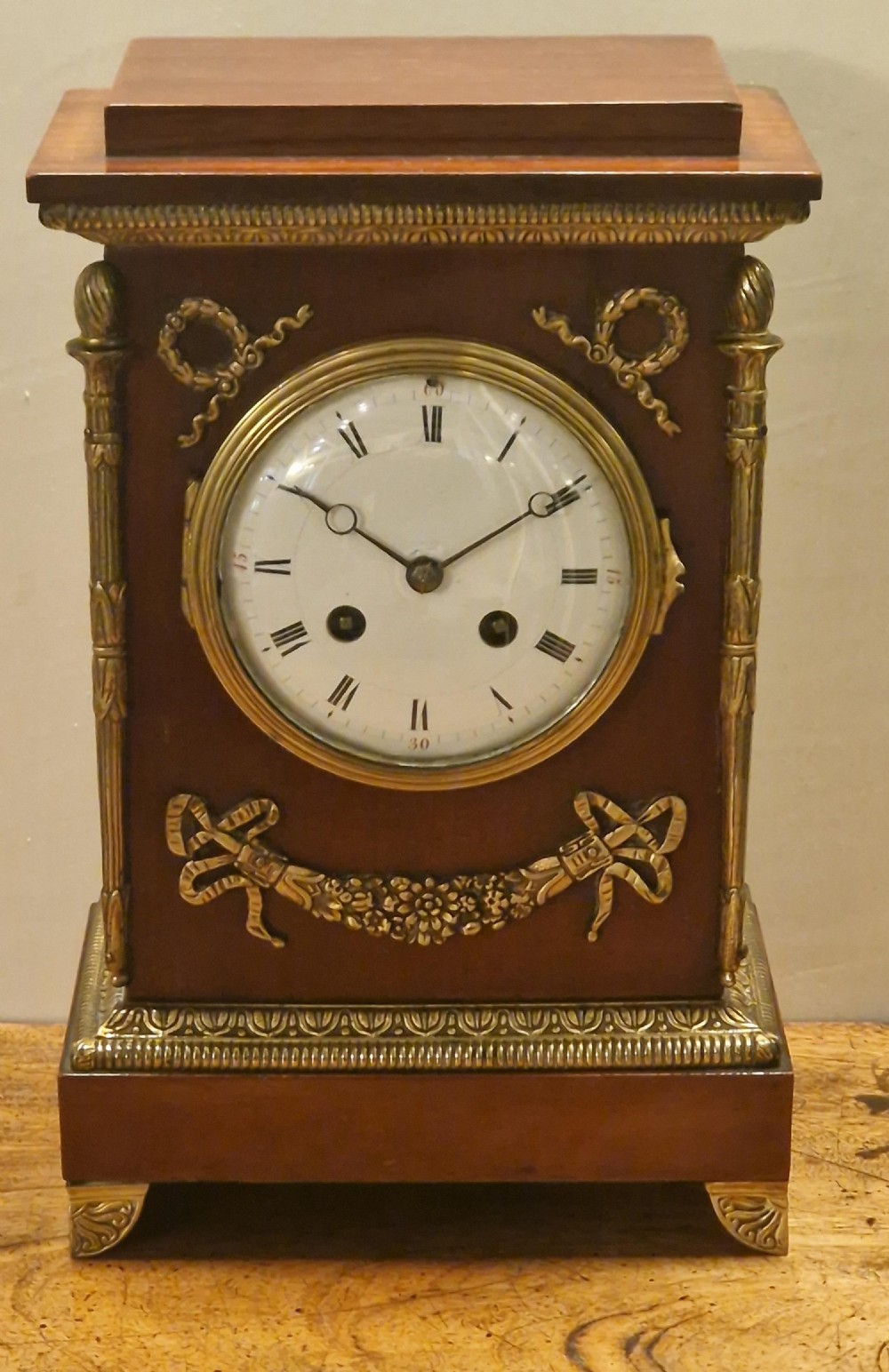 French Empire style mahogany mantel clock featuring gilt-brass floral garlands, fluted corner columns, and a white enamel dial with Roman numerals.