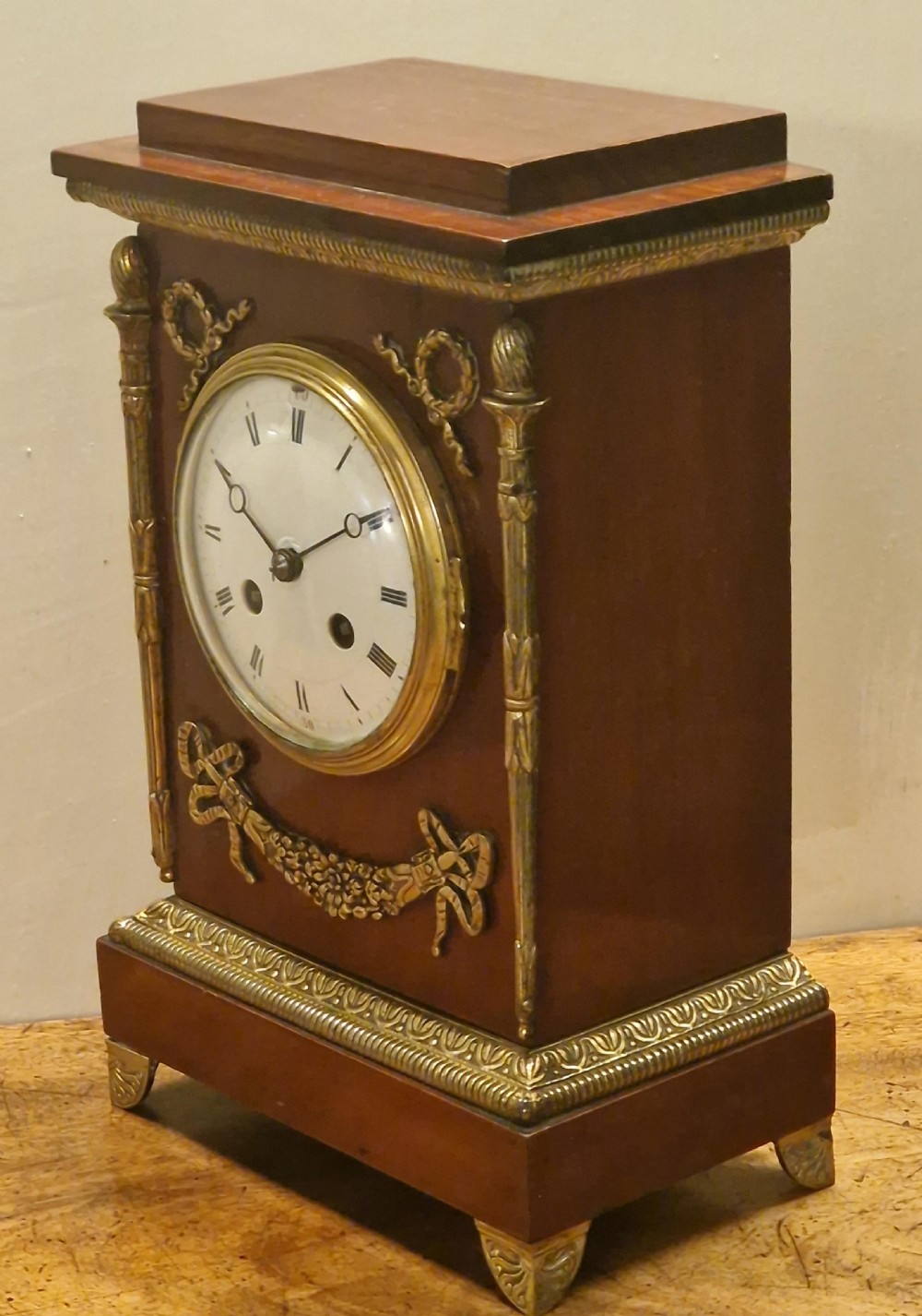 French Empire style mahogany mantel clock featuring gilt-brass floral garlands, fluted corner columns, and a white enamel dial with Roman numerals. View VI