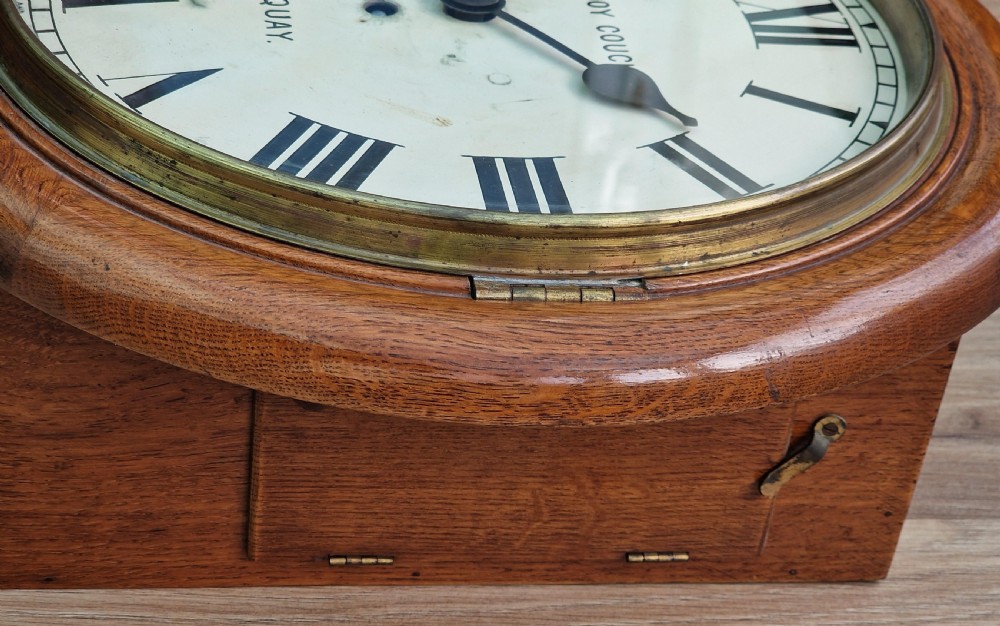 A circular mahogany wall clock with a white painted dial featuring black Roman numerals and black metal hands behind a glass bezel. View V