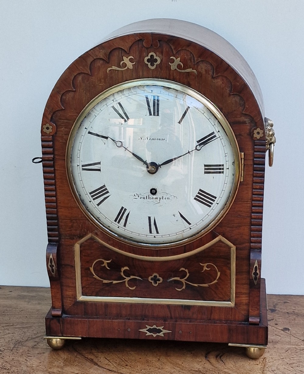 An arched rosewood bracket clock with brass floral inlay, white circular dial signed S. Simpson Southampton with Roman numerals, and side drop handles.