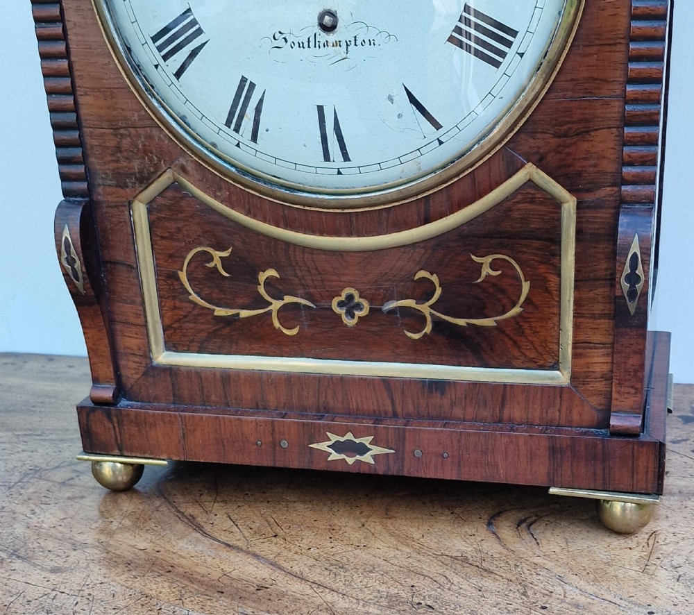 An arched rosewood bracket clock with brass floral inlay, white circular dial signed S. Simpson Southampton with Roman numerals, and side drop handles. View IV