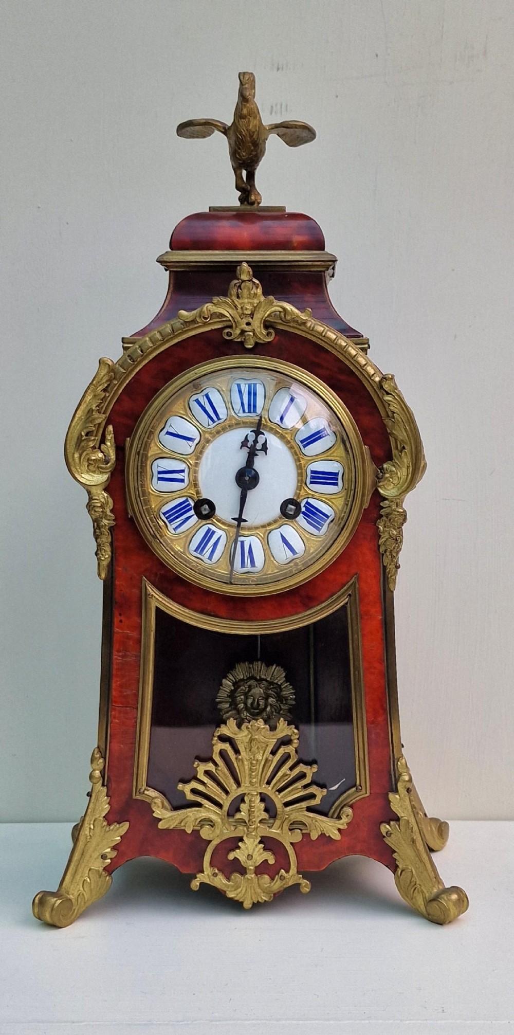 A French Louis XV revival mantel clock featuring red tortoiseshell veneer, gilt bronze ormolu mounts, a top bird finial, and an enamel cartouche dial with blue Roman numerals.