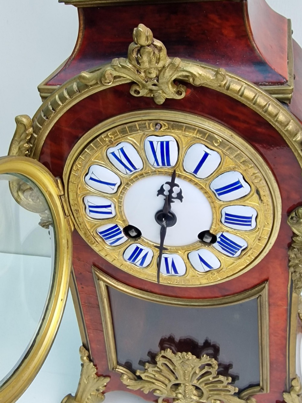 A French Louis XV revival mantel clock featuring red tortoiseshell veneer, gilt bronze ormolu mounts, a top bird finial, and an enamel cartouche dial with blue Roman numerals. View III