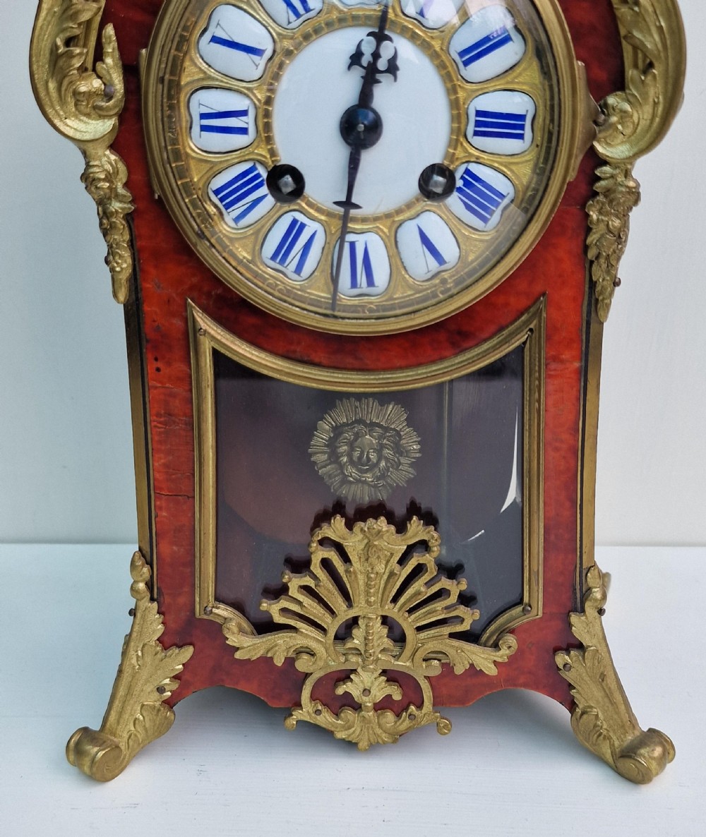 A French Louis XV revival mantel clock featuring red tortoiseshell veneer, gilt bronze ormolu mounts, a top bird finial, and an enamel cartouche dial with blue Roman numerals. View IV