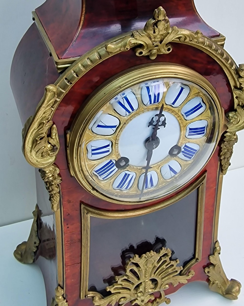 A French Louis XV revival mantel clock featuring red tortoiseshell veneer, gilt bronze ormolu mounts, a top bird finial, and an enamel cartouche dial with blue Roman numerals. View V