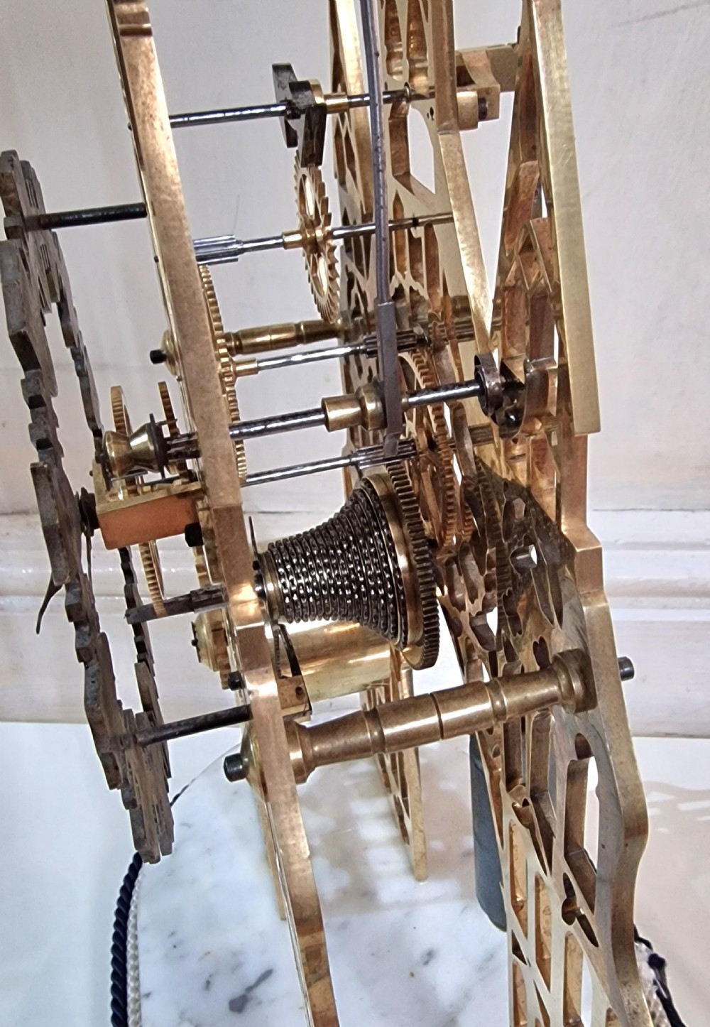 A Victorian Gothic revival brass skeleton clock featuring pierced cathedral spires, a silvered Roman numeral dial, and a top bell, housed under a clear glass dome on a white marble plinth. View V