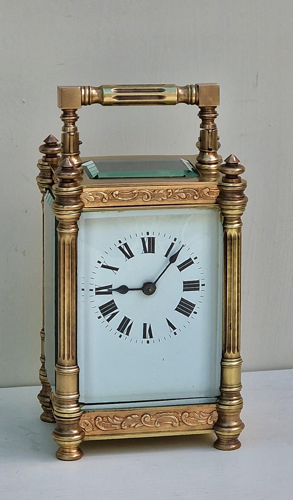 An antique brass carriage clock featuring bevelled glass panels, fluted corner columns, ornate foliage borders, and a white dial with Roman numerals.