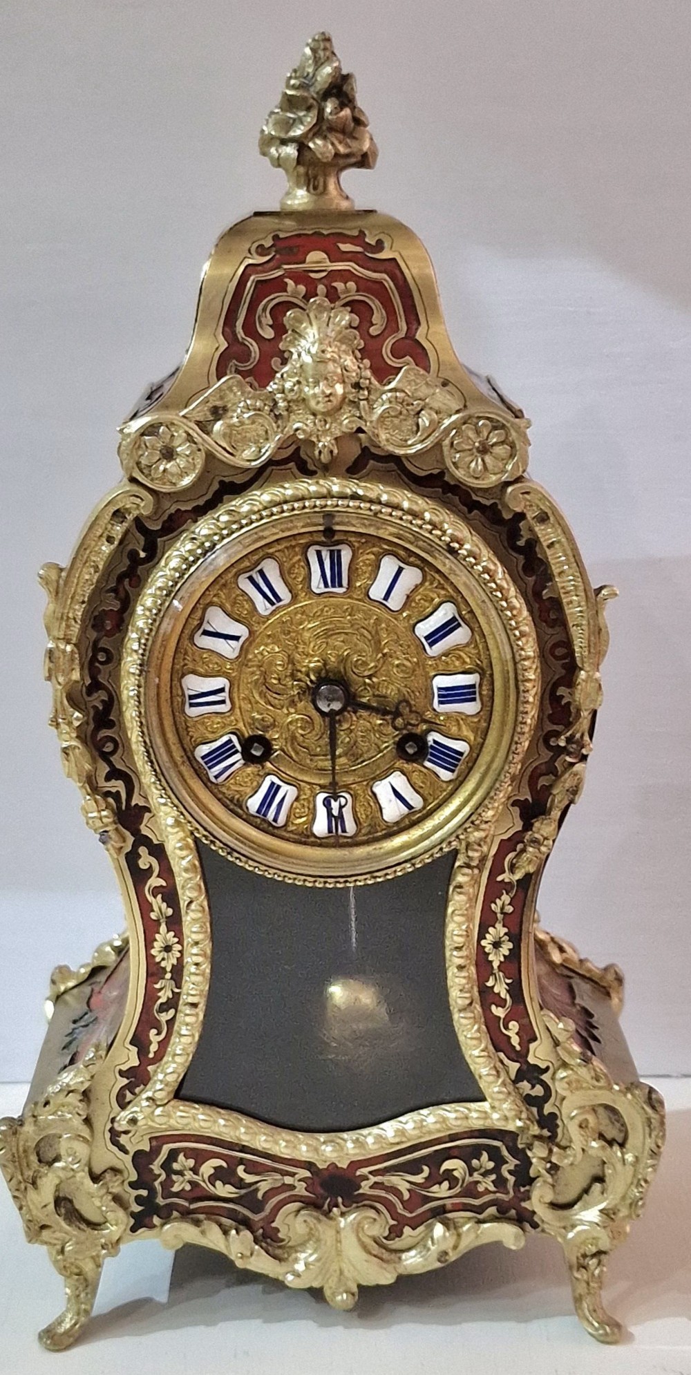 An ornate French Rococo style mantel clock featuring brass and red tortoiseshell Boulle marquetry, gilded ormolu mounts, and a dial with blue Roman numerals on white enamel cartouches.