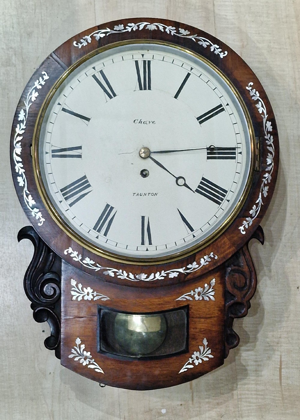 A Victorian drop-dial fusee wall clock featuring a white dial with Roman numerals, a dark wood case adorned with mother-of-pearl floral inlay, and a brass pendulum visible through a glass window.