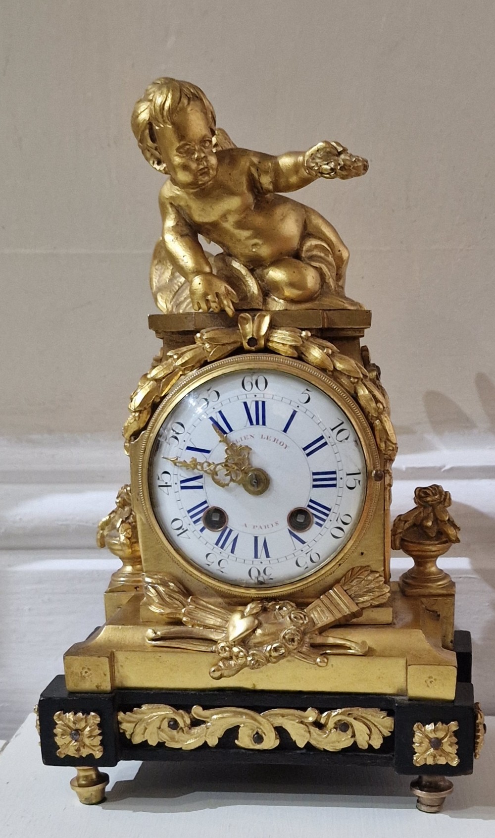 An antique French gilt bronze mantel clock surmounted by a cherub figure resting above a circular white enamel dial with Roman numerals on an ebonised base.