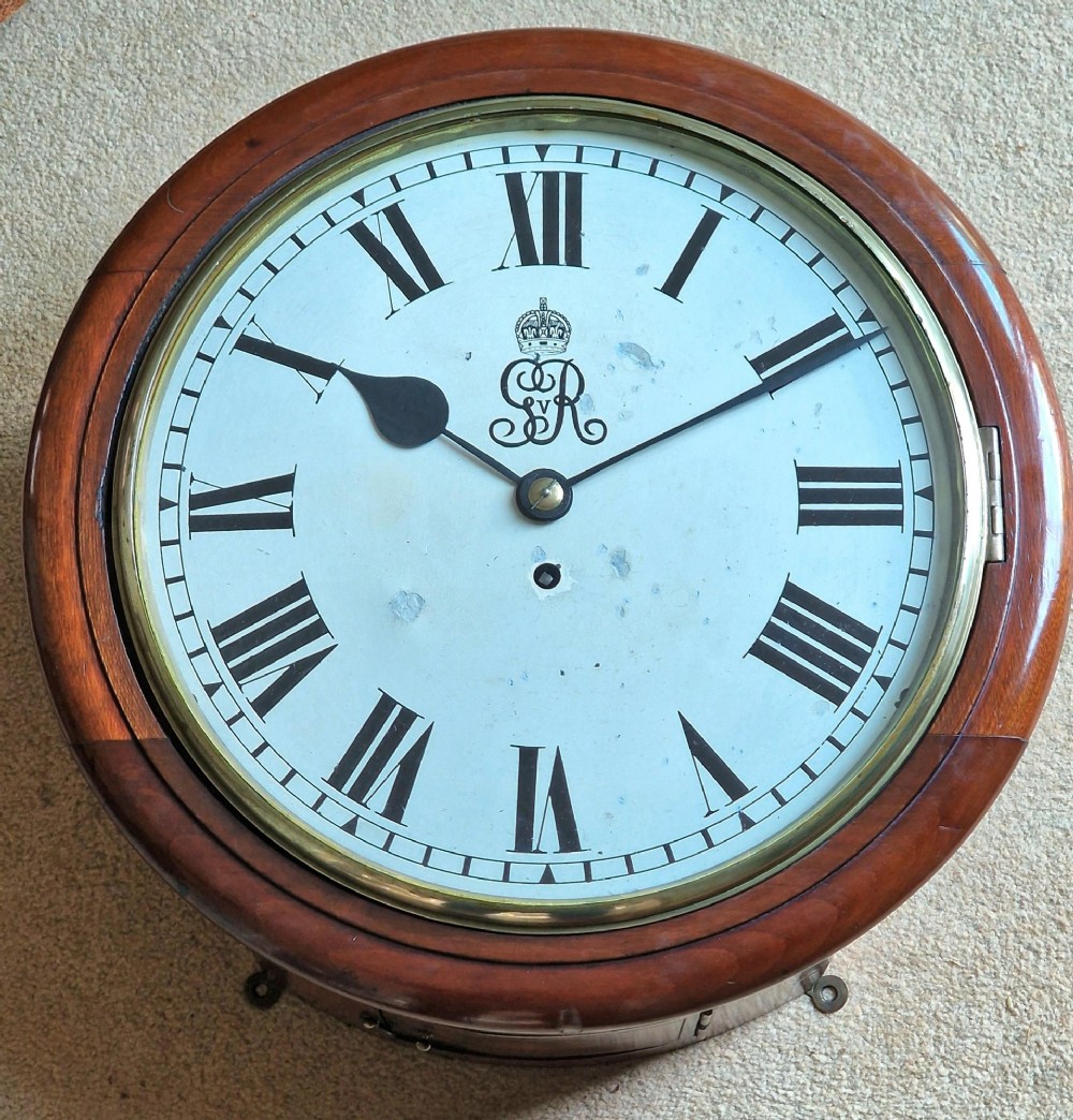 A circular mahogany wall clock with a brass bezel, white painted dial displaying black Roman numerals, and a royal cypher under the twelve oclock position.