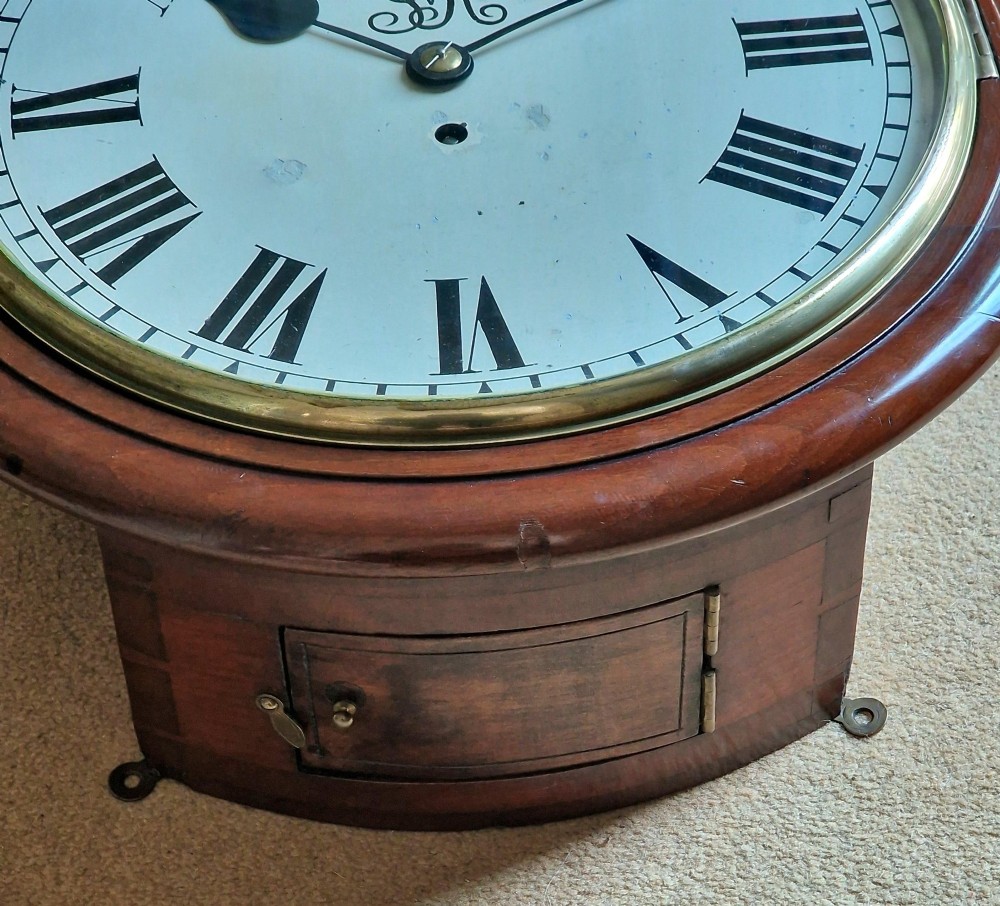A circular mahogany wall clock with a brass bezel, white painted dial displaying black Roman numerals, and a royal cypher under the twelve oclock position. View II