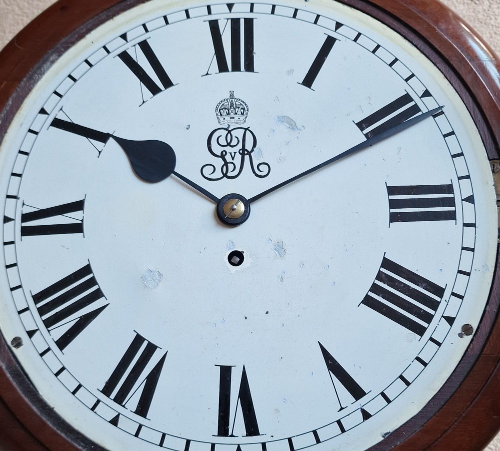 A circular mahogany wall clock with a brass bezel, white painted dial displaying black Roman numerals, and a royal cypher under the twelve oclock position. View III