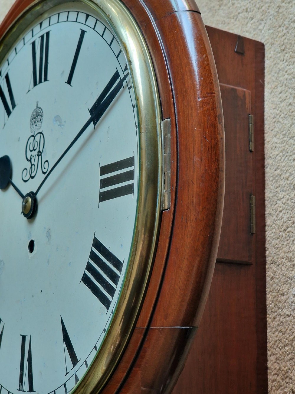 A circular mahogany wall clock with a brass bezel, white painted dial displaying black Roman numerals, and a royal cypher under the twelve oclock position. View VII