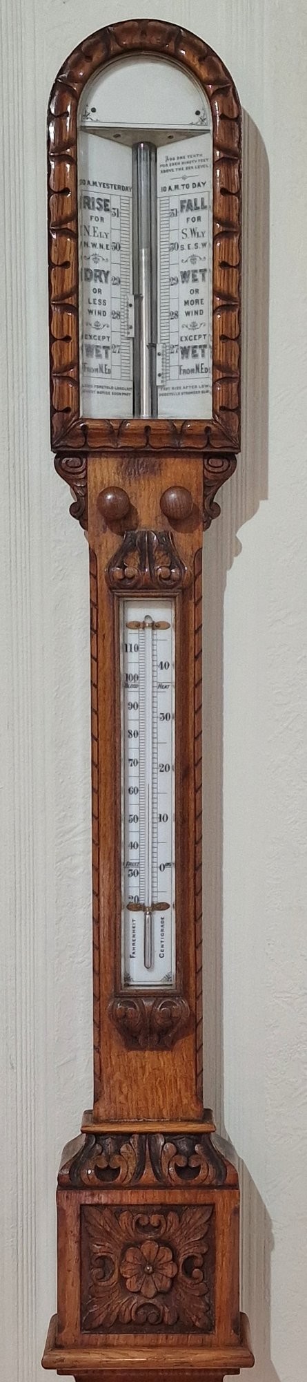 A carved Victorian oak stick barometer featuring an exposed mercury tube, dual weather scales, integrated thermometer, and a carved foliate cistern cover.