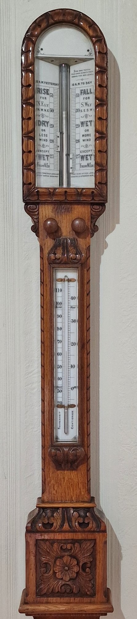 A carved Victorian oak stick barometer featuring an exposed mercury tube, dual weather scales, integrated thermometer, and a carved foliate cistern cover. View V