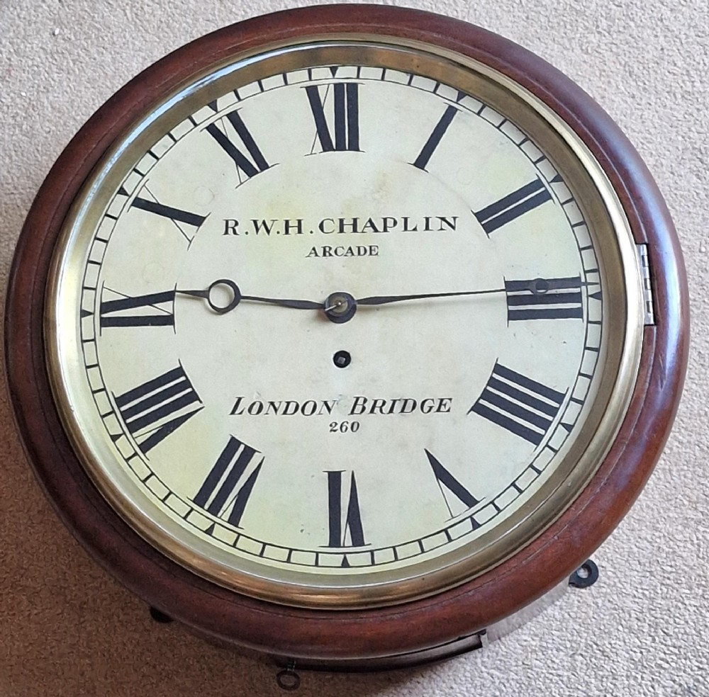 A circular antique mahogany wall clock featuring a cream dial with black Roman numerals and a hinged brass bezel.