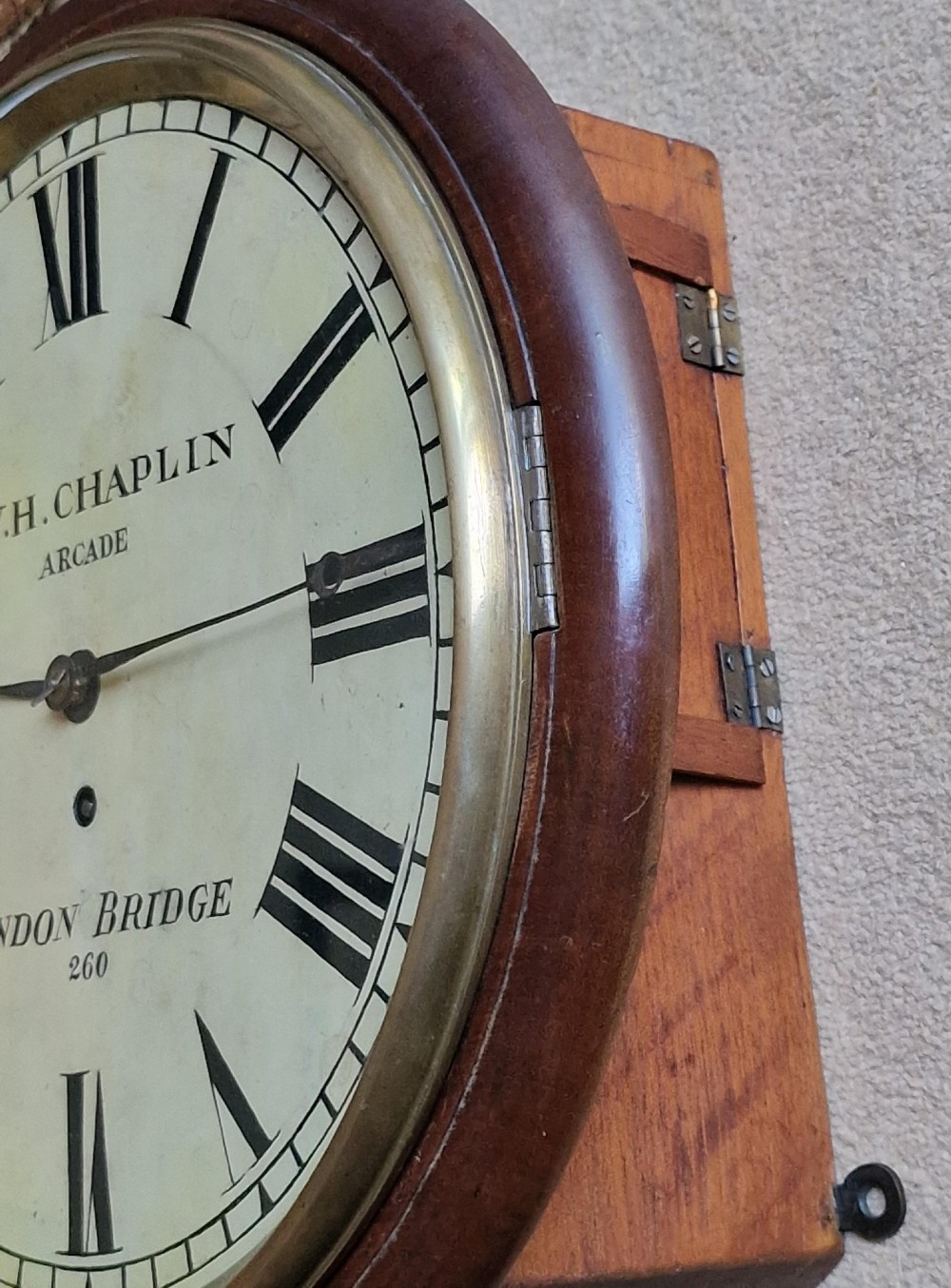 A circular antique mahogany wall clock featuring a cream dial with black Roman numerals and a hinged brass bezel. View IV