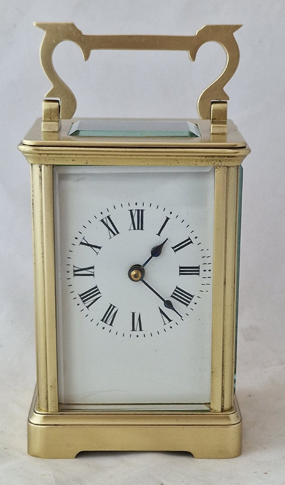 A rectangular brass carriage clock featuring a white enamel dial with Roman numerals, blued steel hands, bevelled glass panels, and a hinged carrying handle on top.
