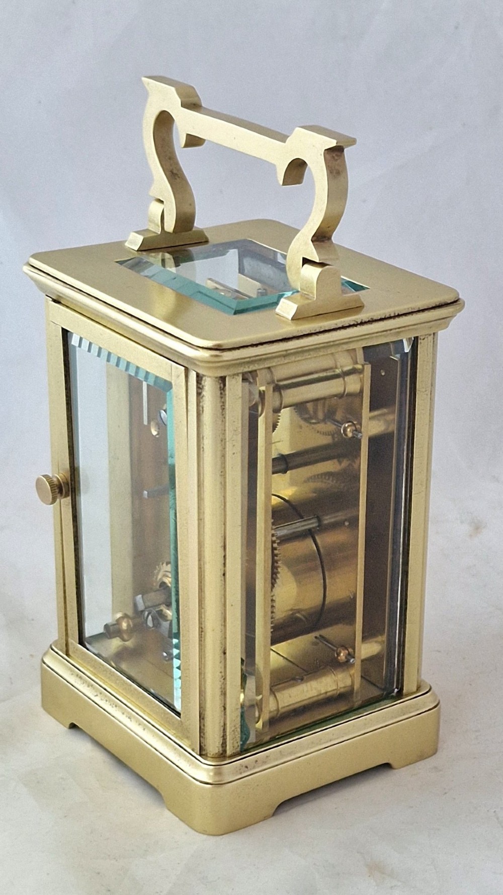 A rectangular brass carriage clock featuring a white enamel dial with Roman numerals, blued steel hands, bevelled glass panels, and a hinged carrying handle on top. View III