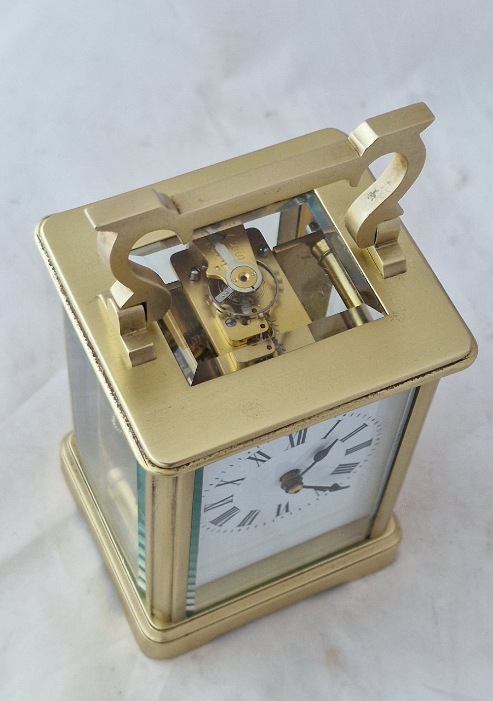 A rectangular brass carriage clock featuring a white enamel dial with Roman numerals, blued steel hands, bevelled glass panels, and a hinged carrying handle on top. View IV