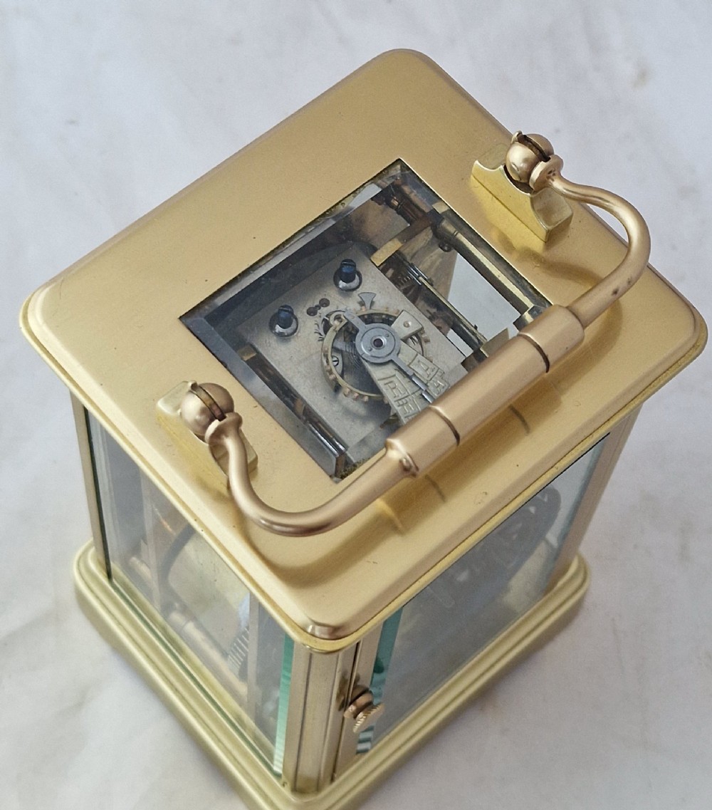 A brass carriage clock featuring bevelled glass side panels, a white enamel dial with black Roman numerals, and a top carrying handle. View III