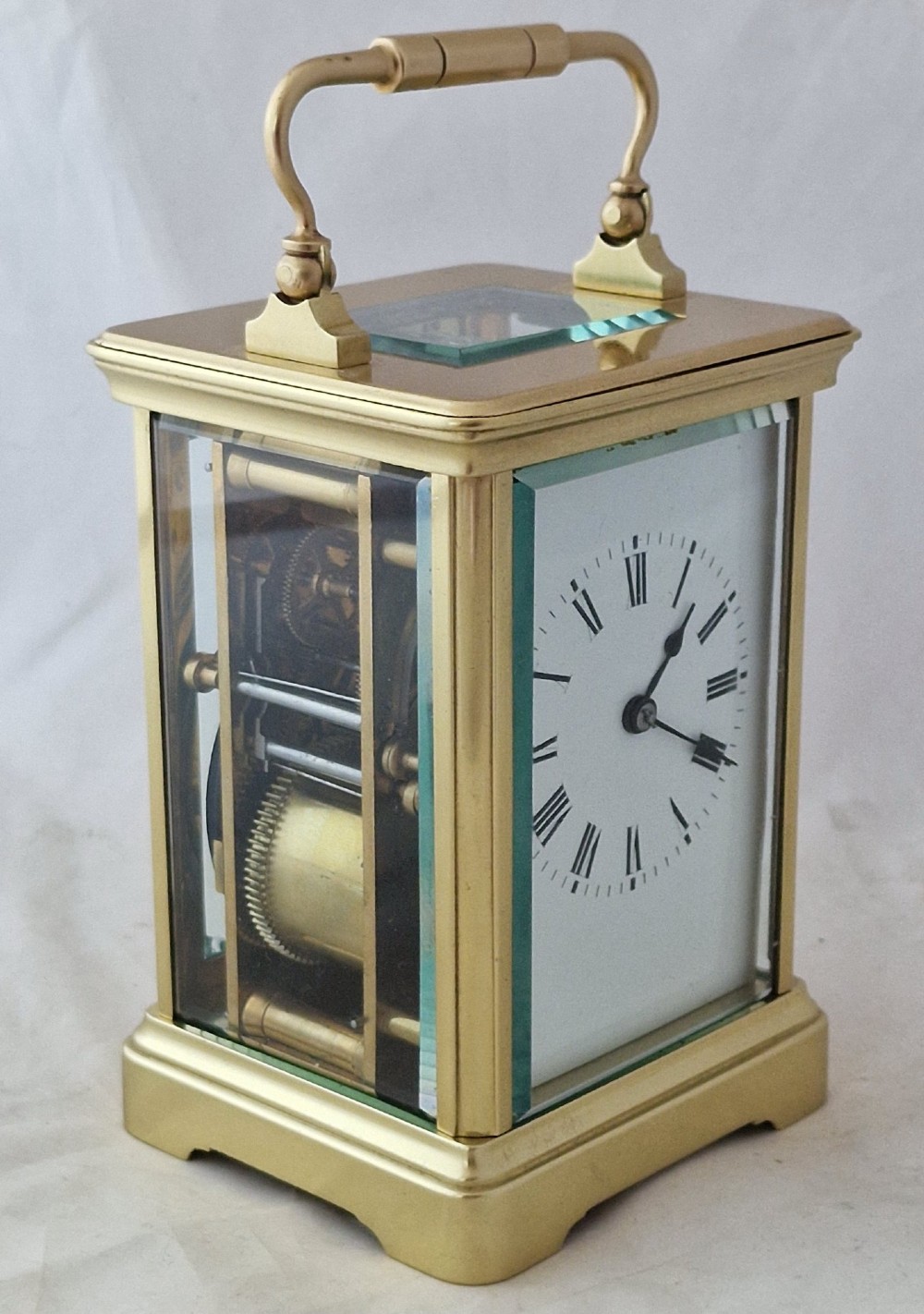 A brass carriage clock featuring bevelled glass side panels, a white enamel dial with black Roman numerals, and a top carrying handle. View IV