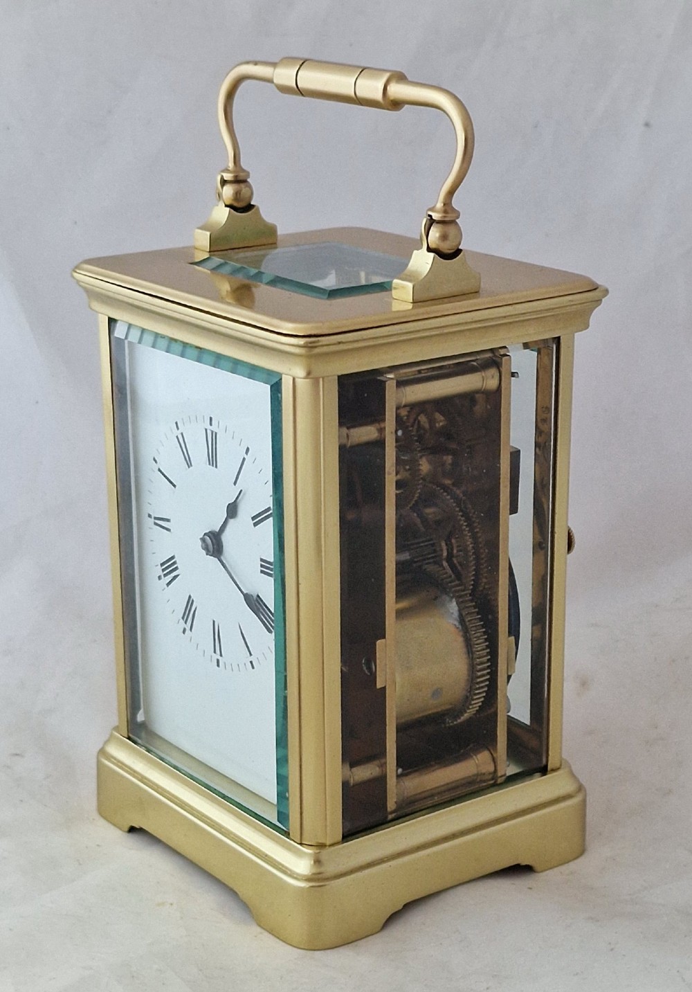 A brass carriage clock featuring bevelled glass side panels, a white enamel dial with black Roman numerals, and a top carrying handle. View V