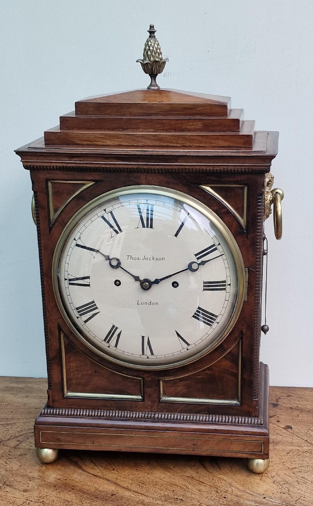 A Regency mahogany bracket clock featuring a white circular dial signed Thomas Jackson London, framed by brass stringing, lion head side handles, and a pineapple finial.