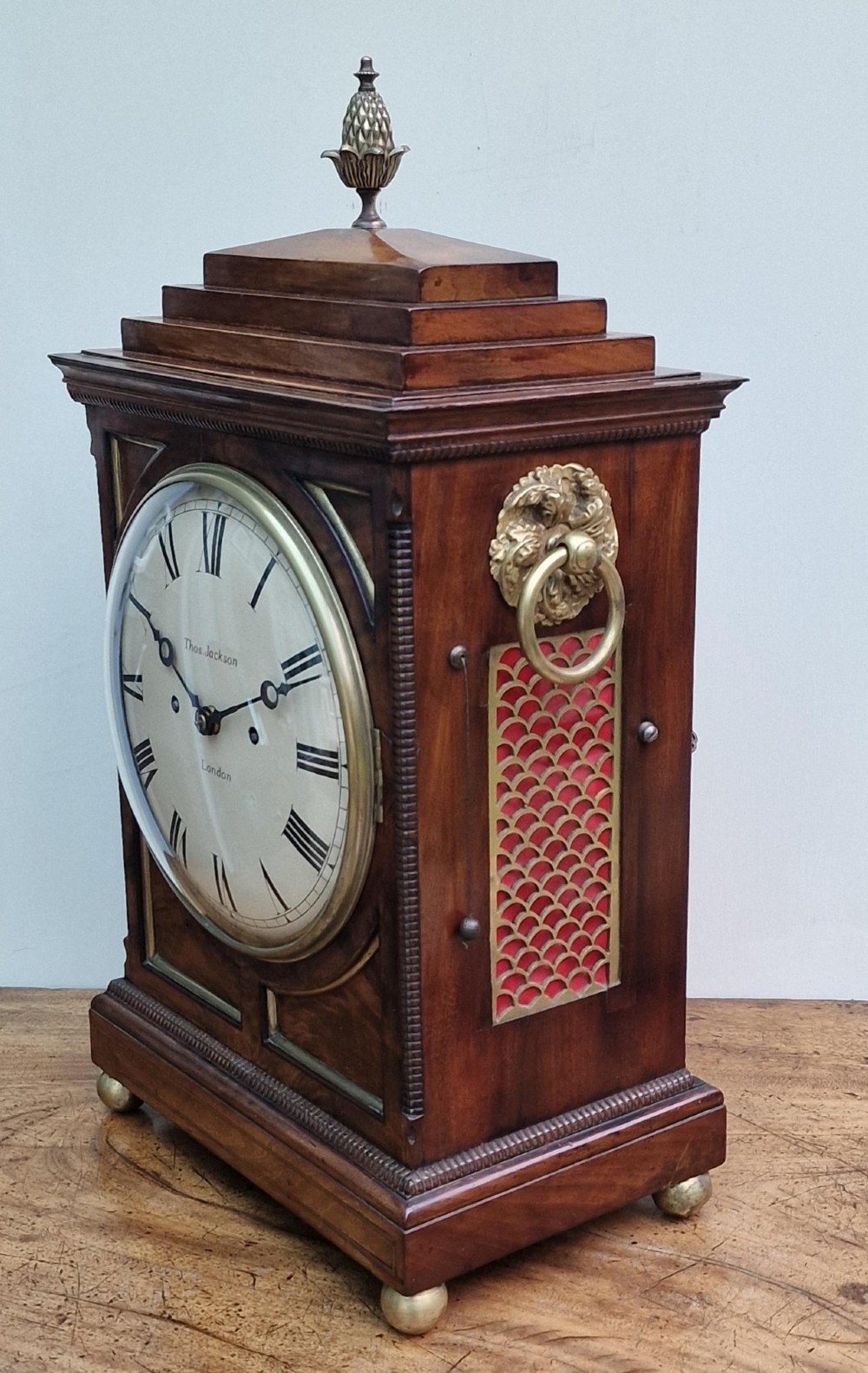 A Regency mahogany bracket clock featuring a white circular dial signed Thomas Jackson London, framed by brass stringing, lion head side handles, and a pineapple finial. View V