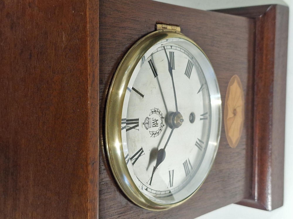 An upright wooden mantel clock featuring a brass-rimmed dial marked with the RAF crest and Roman numerals, set above an oval floral marquetry inlay on a stepped base. View VI