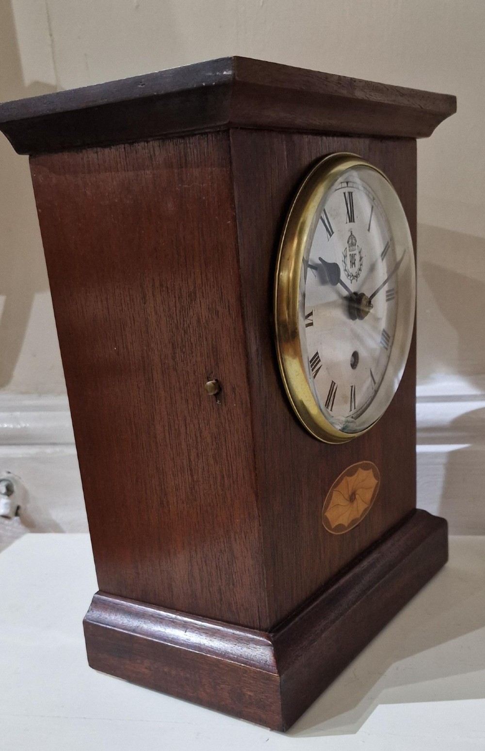 An upright wooden mantel clock featuring a brass-rimmed dial marked with the RAF crest and Roman numerals, set above an oval floral marquetry inlay on a stepped base. View VII