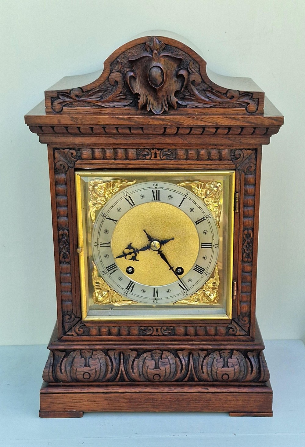 A carved oak mantel clock featuring an arched pediment, ornate brass dial with a silvered chapter ring, and a carved decorative base.