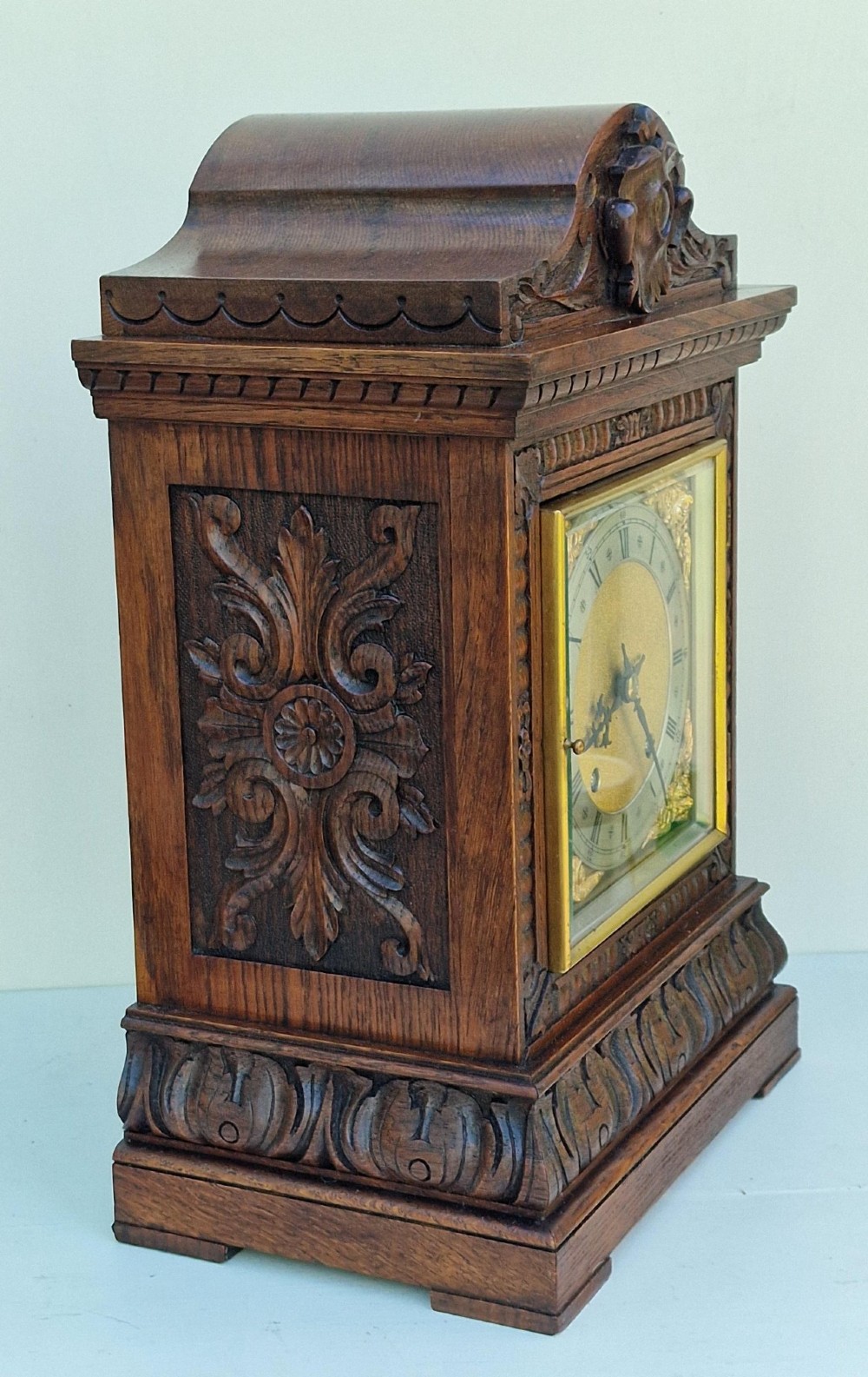 A carved oak mantel clock featuring an arched pediment, ornate brass dial with a silvered chapter ring, and a carved decorative base. View IV