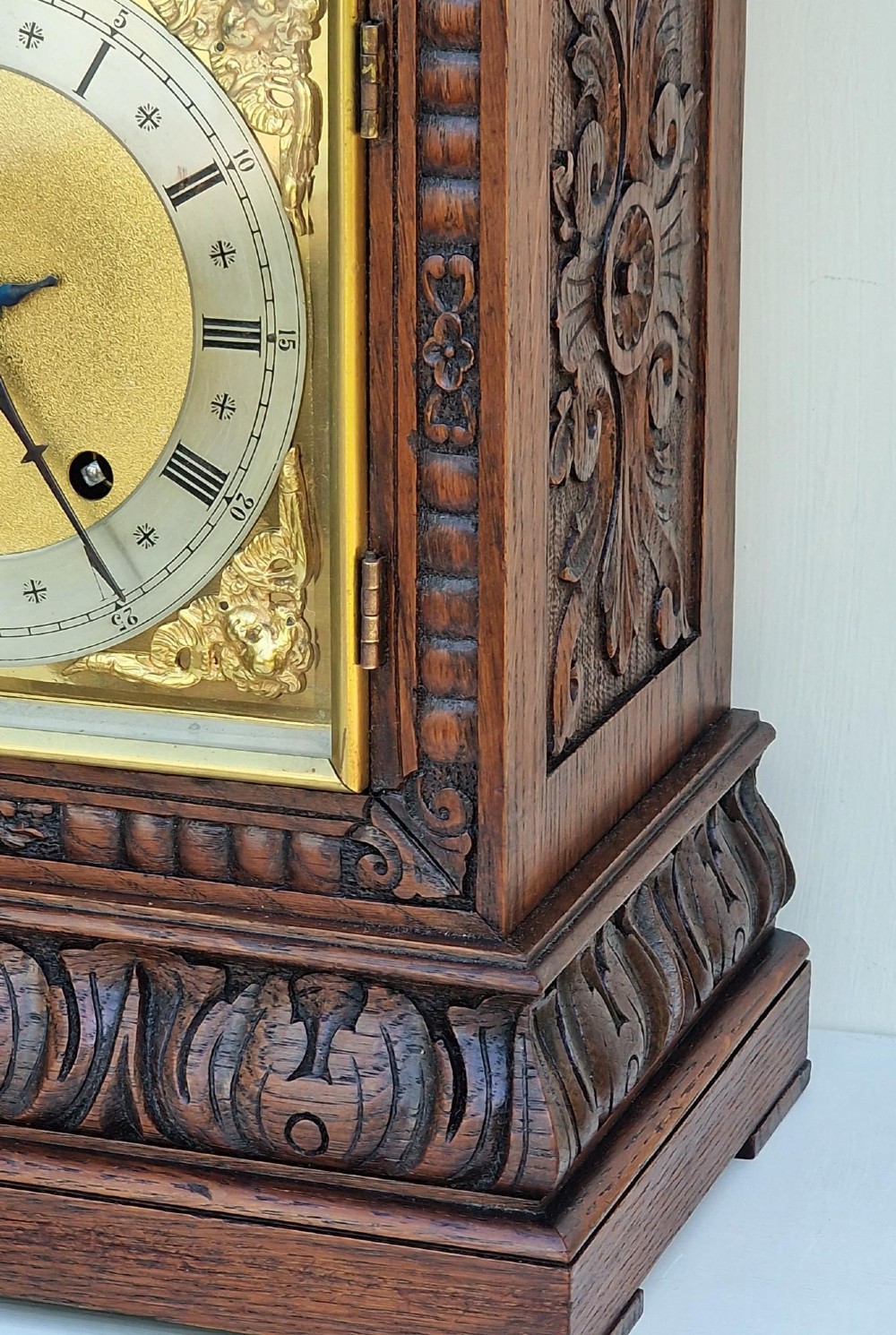 A carved oak mantel clock featuring an arched pediment, ornate brass dial with a silvered chapter ring, and a carved decorative base. View VI